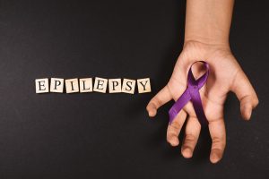 epilepsy spelt with scabble tiles on a black background and someone's hand holding a purple ribbon