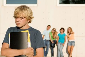 Isolated teenage boy in the forground with fellow 'popular' high school pupils learning against the wall staring on the background.